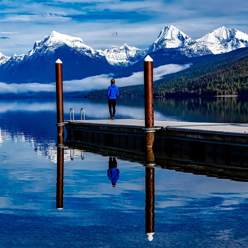 person standing on dock looking out at water and snow covered mountains