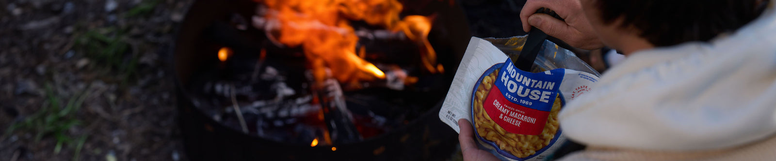 Person sitting by a campfire at dusk enjoying a pouch of Mountain House Vegetarian Creamy Macaroni and Cheese.