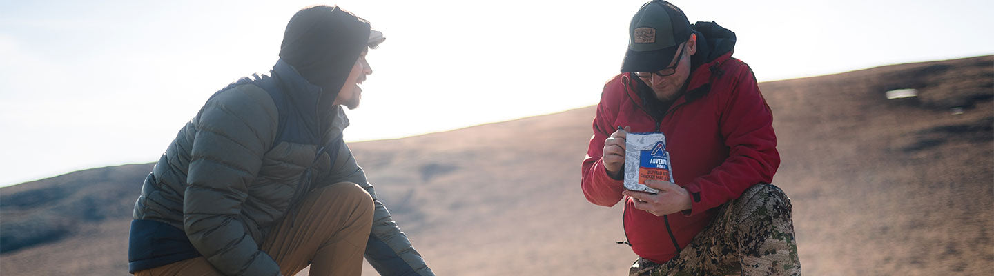 Guy holding looking inside pouch of Mountain House with another person looking at him out in the backcountry