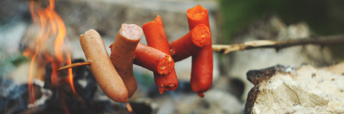 Close-up of multiple sausages cooking on a single stick over a campfire in the wilderness.