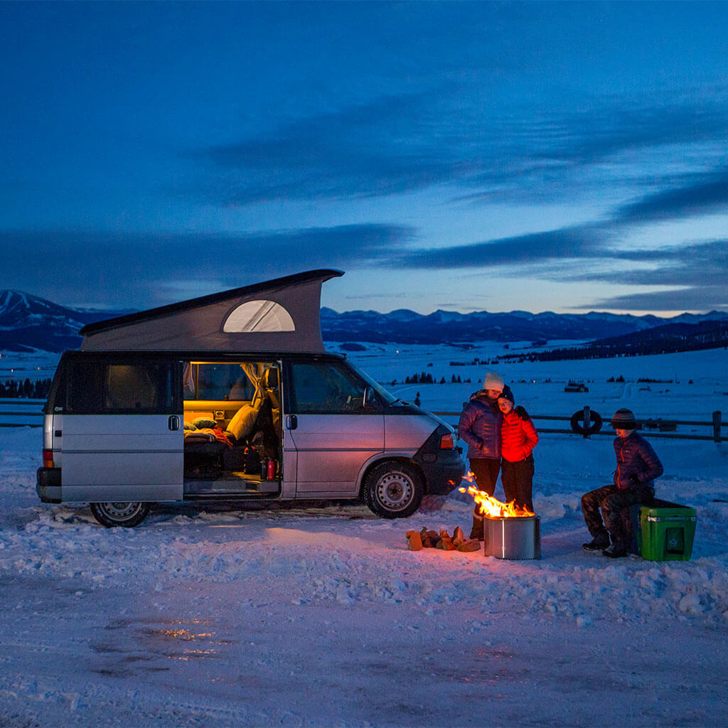 people standing around campfire during winter with camper van in background