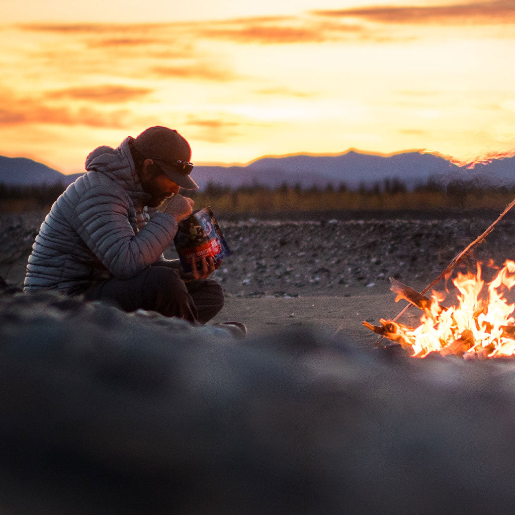Guy eating from Mountain House freeze-dried food pouch by campfire at dusk.