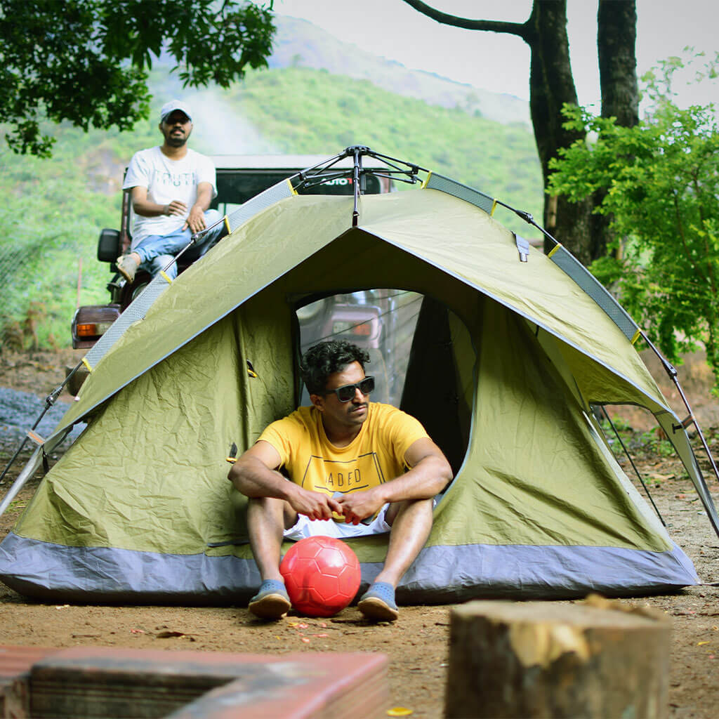 man sitting in tent while other man behinds sits on hood of vehicle