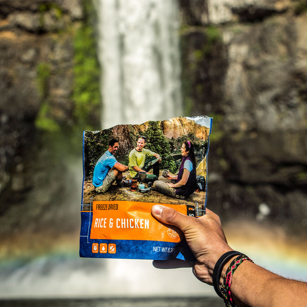 person holding pouch of mountain house freeze dried rice and chicken in front of waterfall