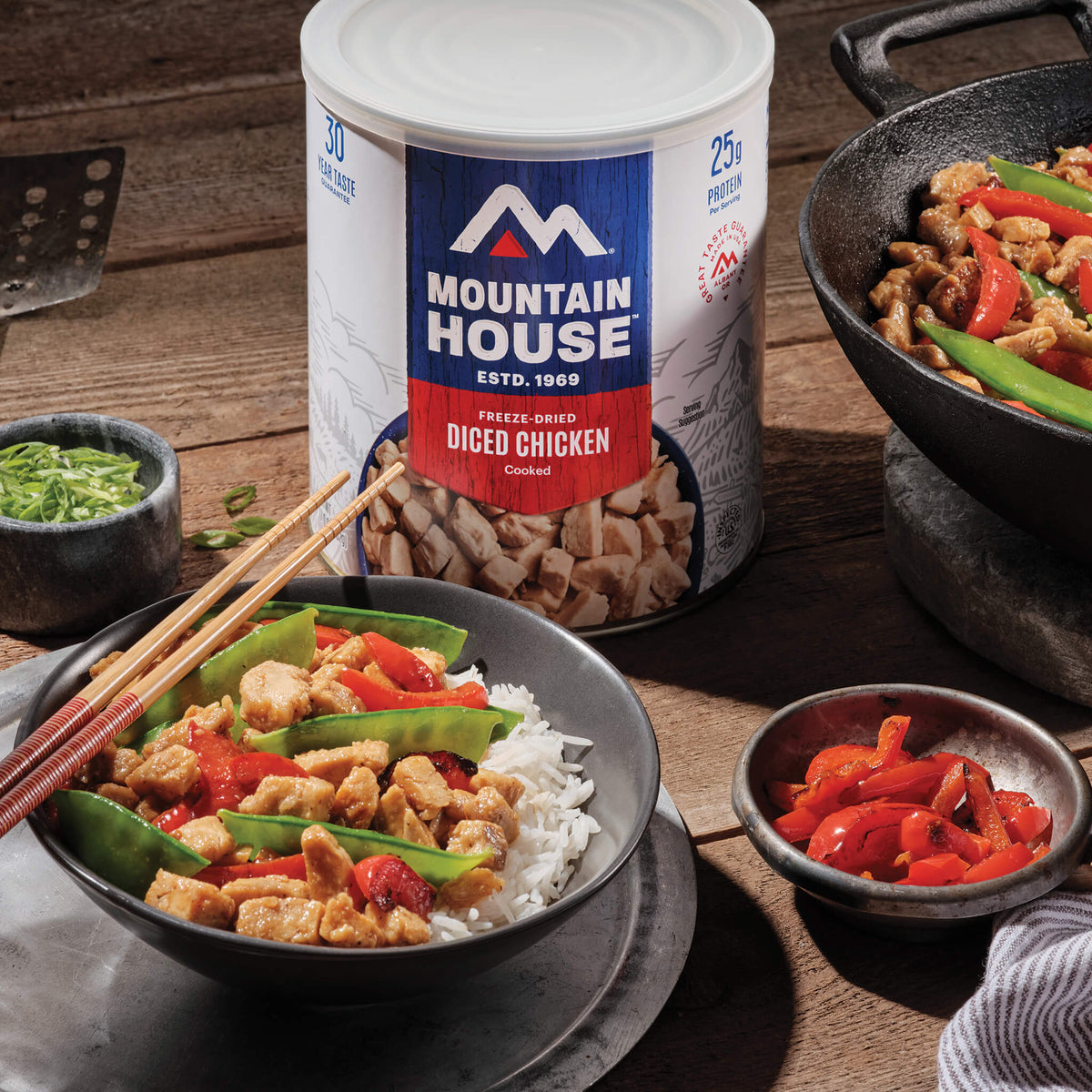Mountain House freeze-dried diced chicken can on a wooden table with a bowl of chicken stir-fry.