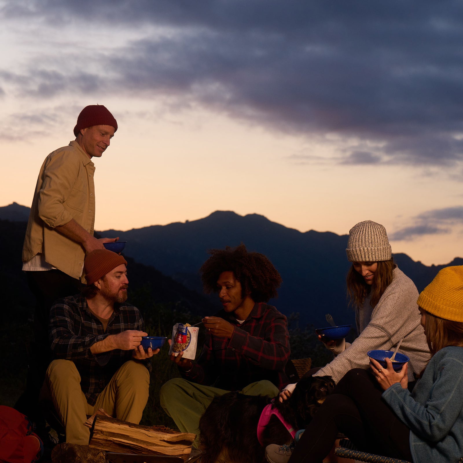 A group of people gathered around a firepit at dusk, enjoying their meal from bowls and a Mountain House pouch of Beef Stroganoff.