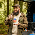 Guy standing by open truck bed with various camping supplies eating from a a pouch of Mountain House Beef Stroganoff with Noodles.