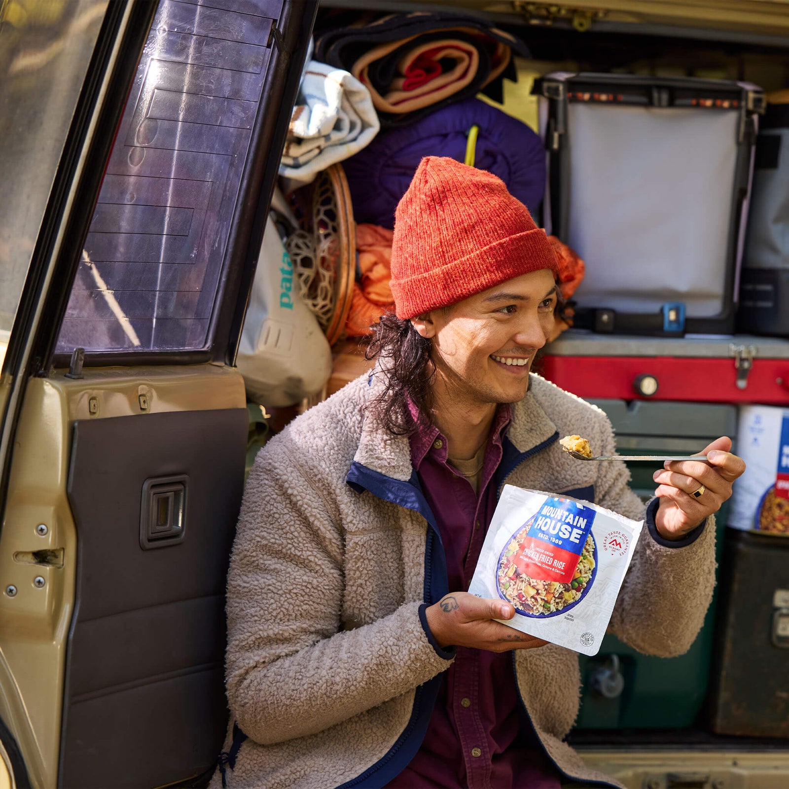 Person in a beanie sitting on bumper of loaded SUV with camping supplies eating from a pouch of Mountain House Chicken Fried Rice.