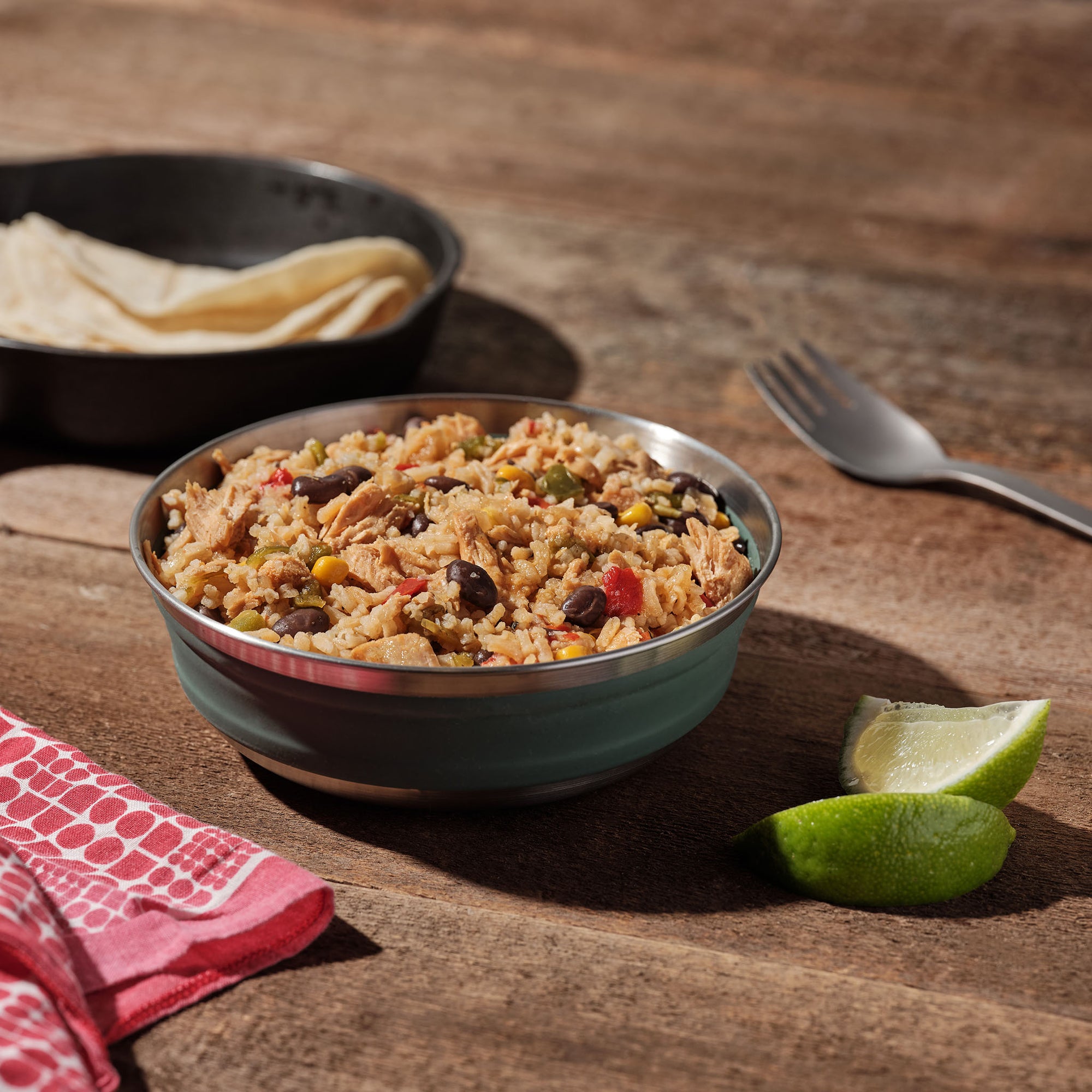 A blue camping bowl filled with Mountain House Chicken Fajita sits on a picnic table, surrounded by a cast iron skillet of tortillas, fresh limes, a camping fork, and a red napkin.