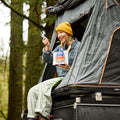 Person sitting at the door of a roof tent eating from a pouch of Mountain House Kung Pao Chicken while camping.