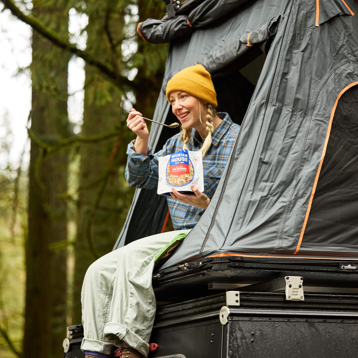 Person sitting at the door of a roof tent eating from a pouch of Mountain House Kung Pao Chicken while camping.