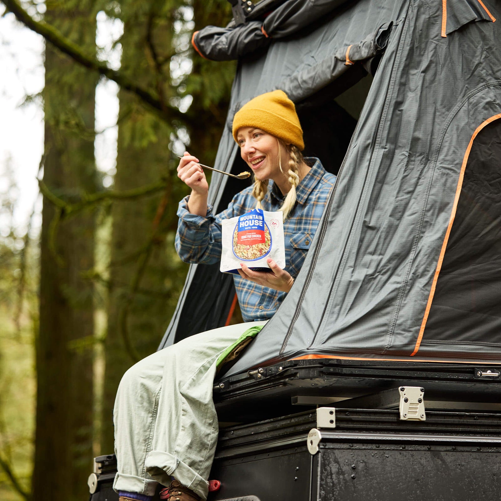Person sitting at the door of a roof tent eating from a pouch of Mountain House Kung Pao Chicken while camping.