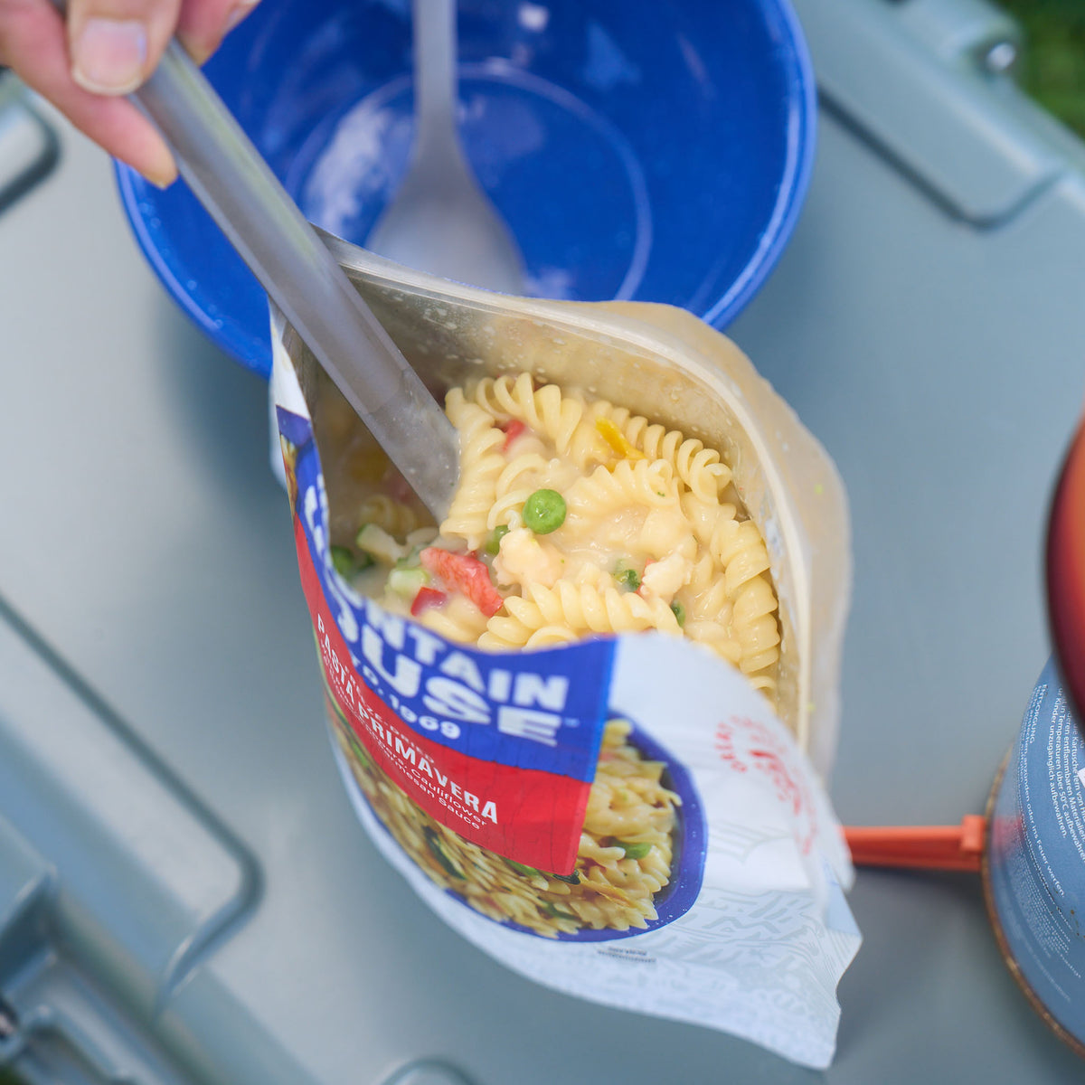 A close-up view of a pouch of Mountain House Pasta Primavera, featuring delicate spiral noodles, green peas, zucchini, and other vegetables in a creamy parmesan cheese sauce. The pouch sits on top of a cooler next to a blue camping bowl and portable stove.