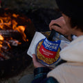 Person sitting by a campfire at night eating from a pouch of Mountain House Creamy Macaroni & Cheese.