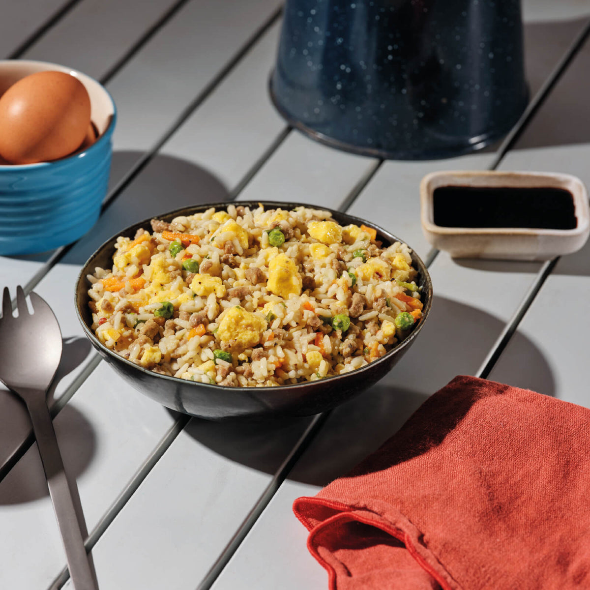 A black bowl filled with Mountain House Breakfast Fried Rice on a camping table with a spork, eggs, soy sauce, and kettle in the background.