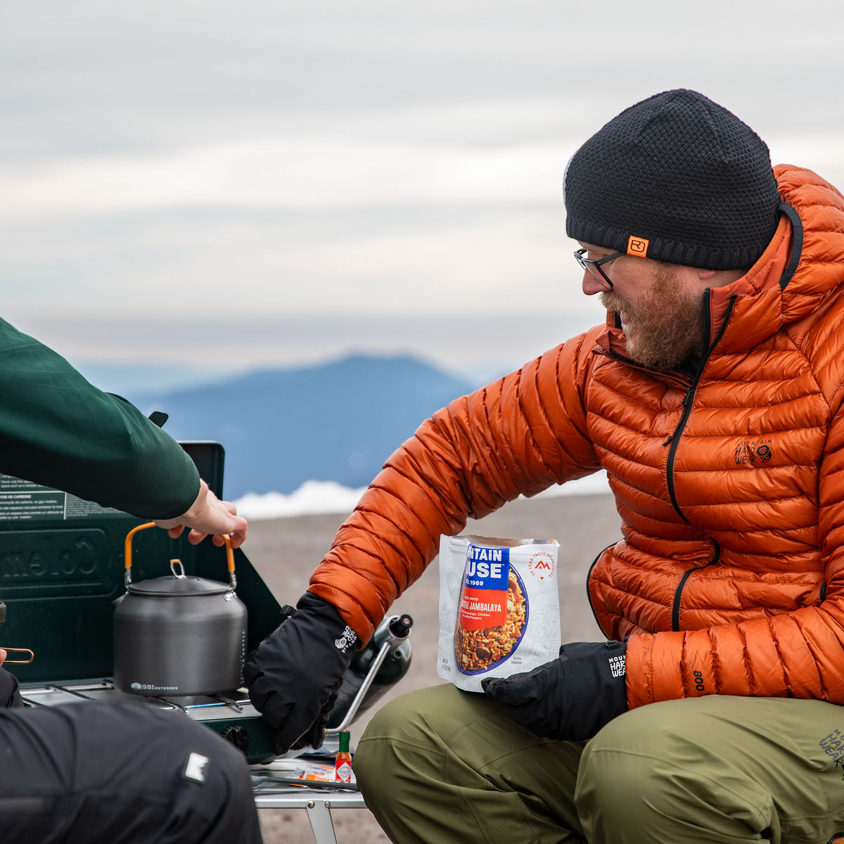 Person in orange jacket sitting outdoors with a camping stove and holding a pouch of Mountain House Cajun Jambalaya with mountains in the background during winter.