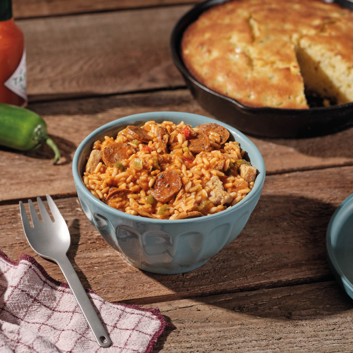 Blue bowl of Cajun Jambalaya with a skillet of cornbread, jalapeno, and hot sauce in the background on a wooden table.