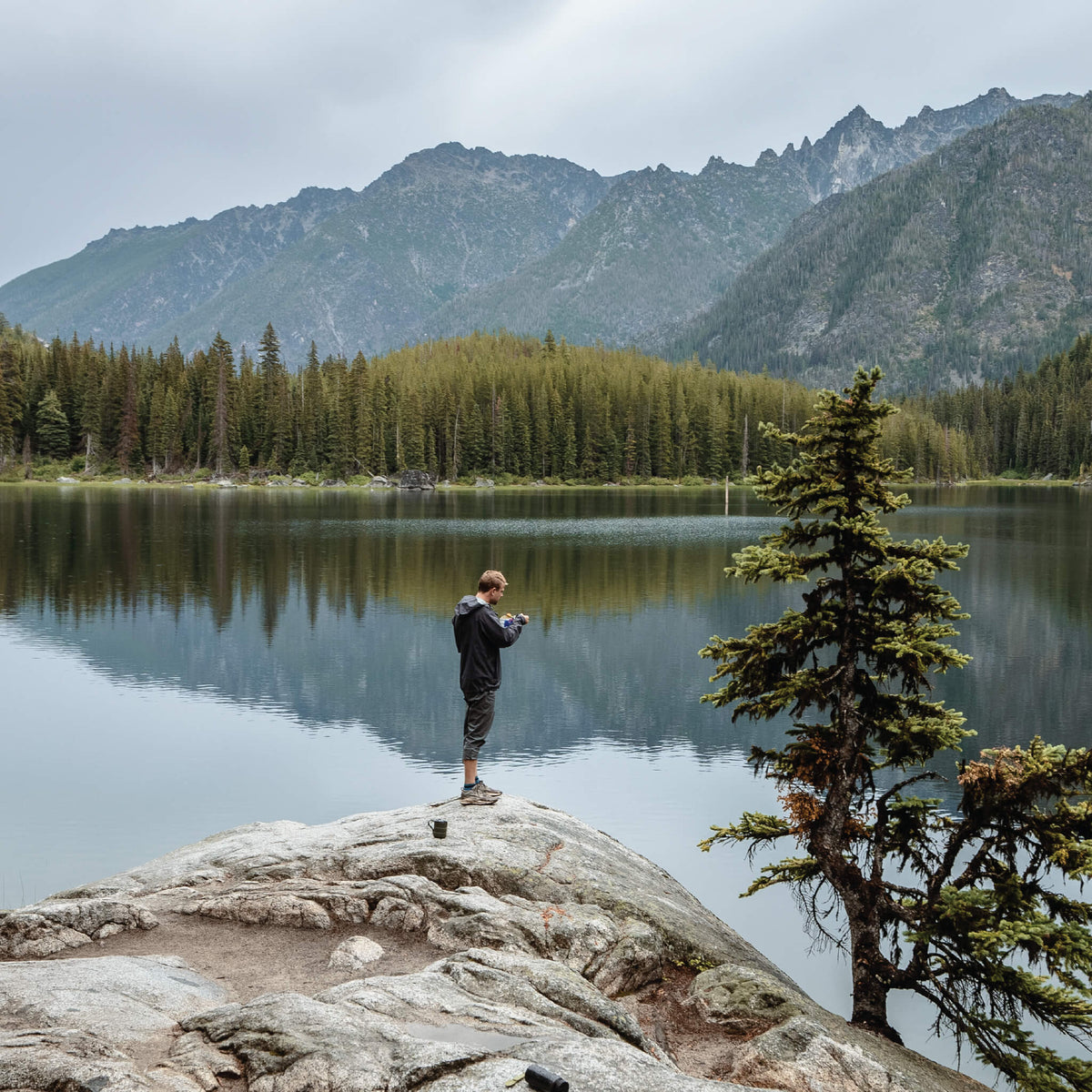 Person eating Mountain House from a pouch on a rocky shoreline next to a lake, with rugged mountains in the background.
