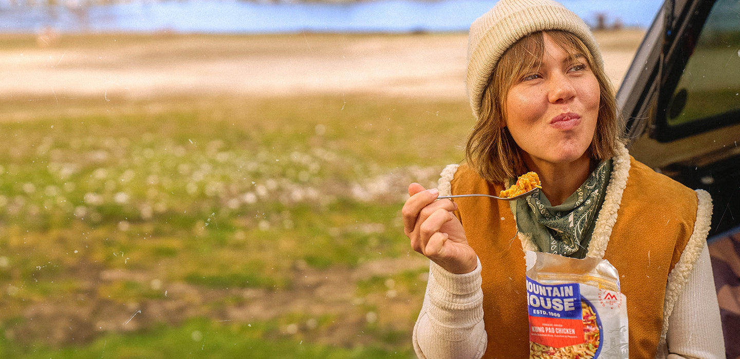 Woman sitting on a tailgate in a field eating a Kung Pao Chicken Mountain House pouch