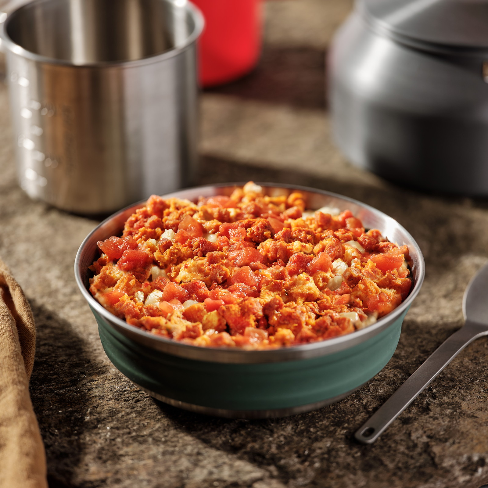 A bowl of Mountain House Cheesy Pepperoni Pizza Bowl in a camping bowl, with various camp kitchen supplies in the background.