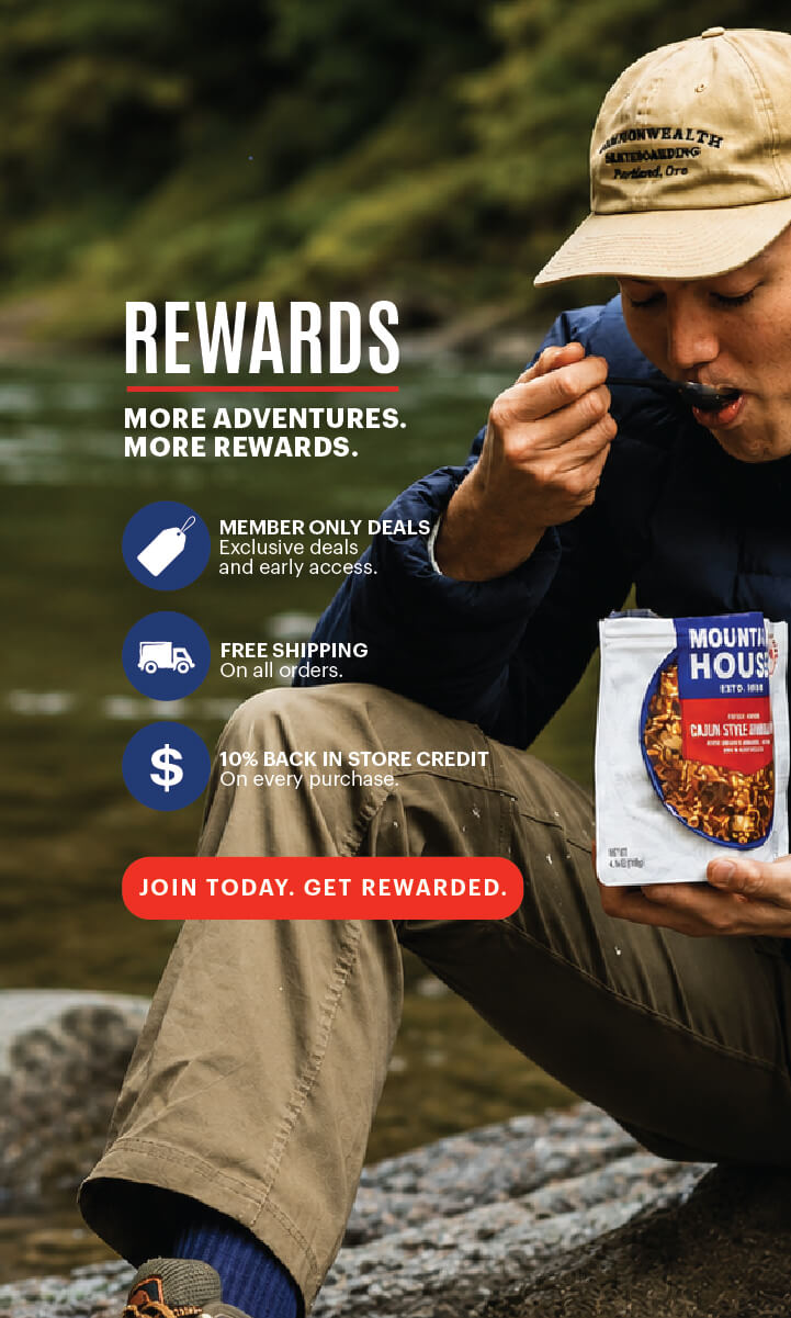 Person eating Mountain House Cajun Style Jambalaya by a river; Rewards program offers deals, free shipping, and credits.
