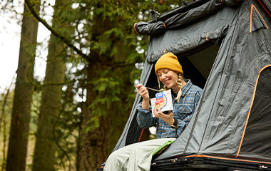 Girl sitting at the doorway of a roof tent while camping eating from a Mountain House pouch.
