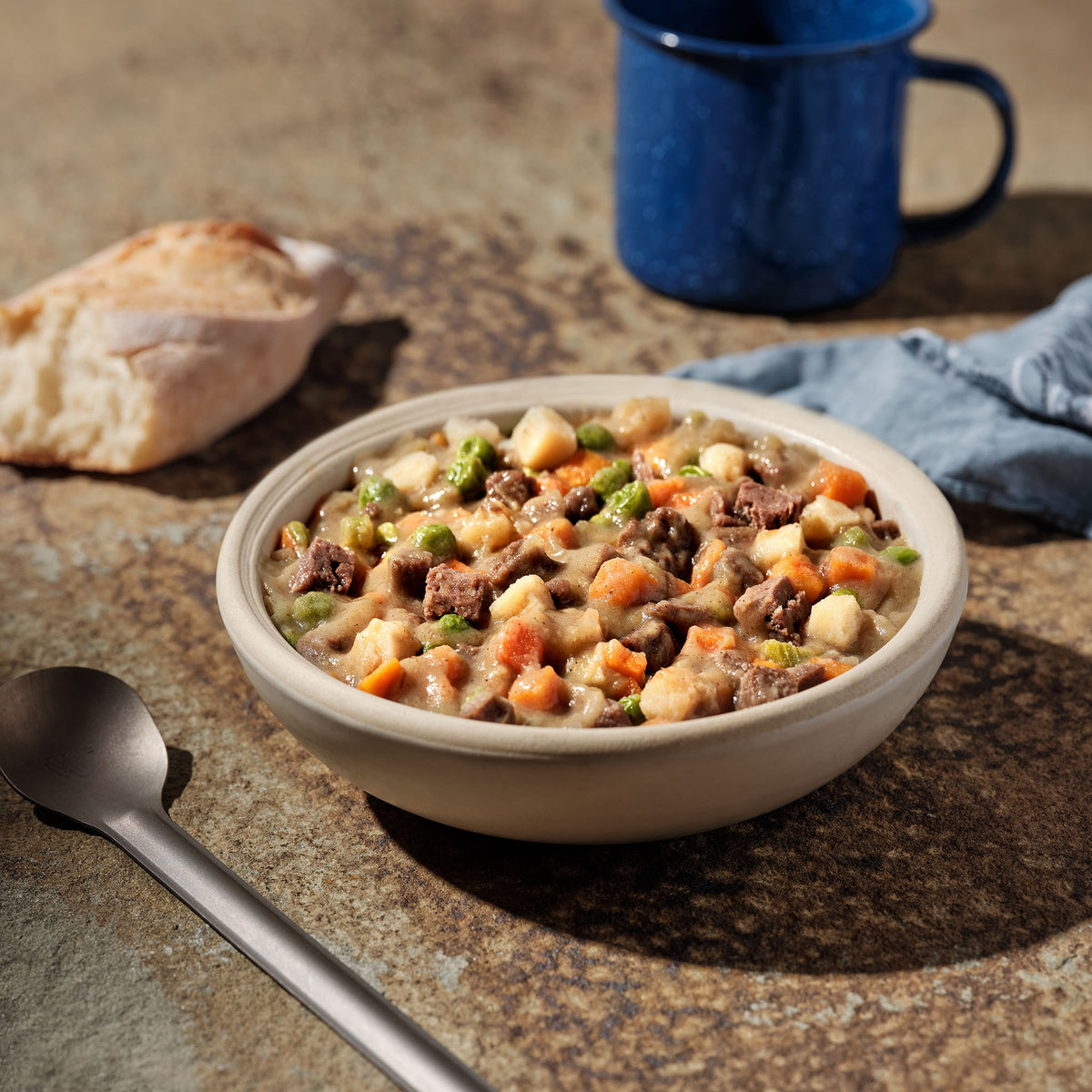 A bowl of Mountain House Beef Stew sits on a counter, surrounded by a blue camping cup, a loaf of bread, a spoon, and a blue napkin.
