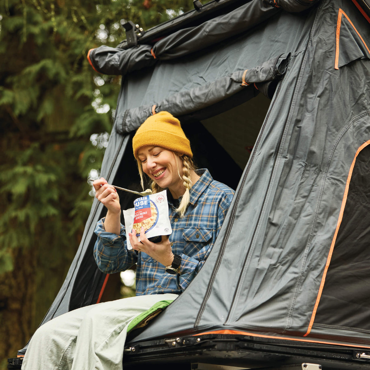Girl sitting inside a rooftop tent eating from a Mountain House pouch in the forest