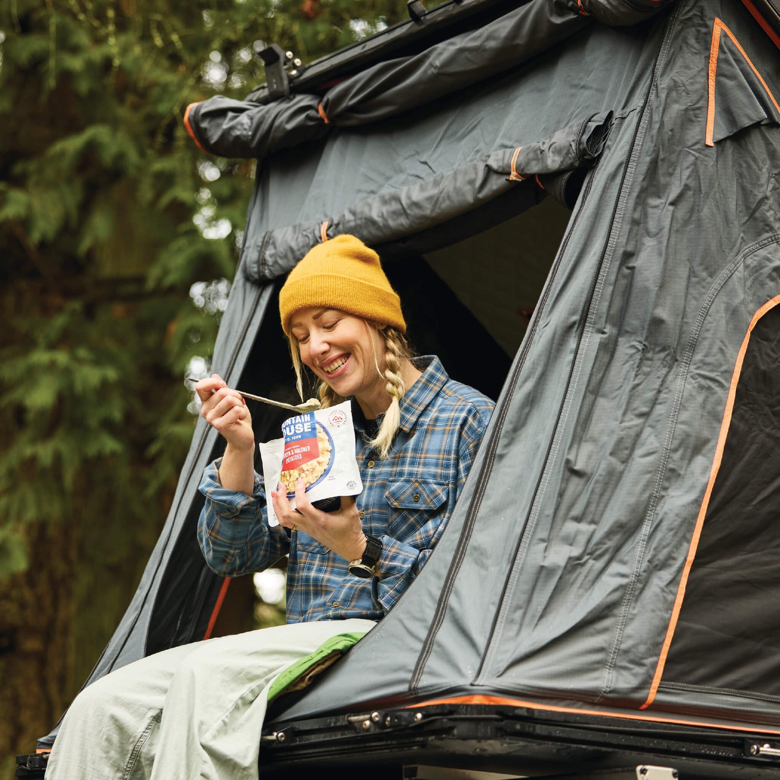 Girl sitting inside a rooftop tent eating from a Mountain House pouch in the forest