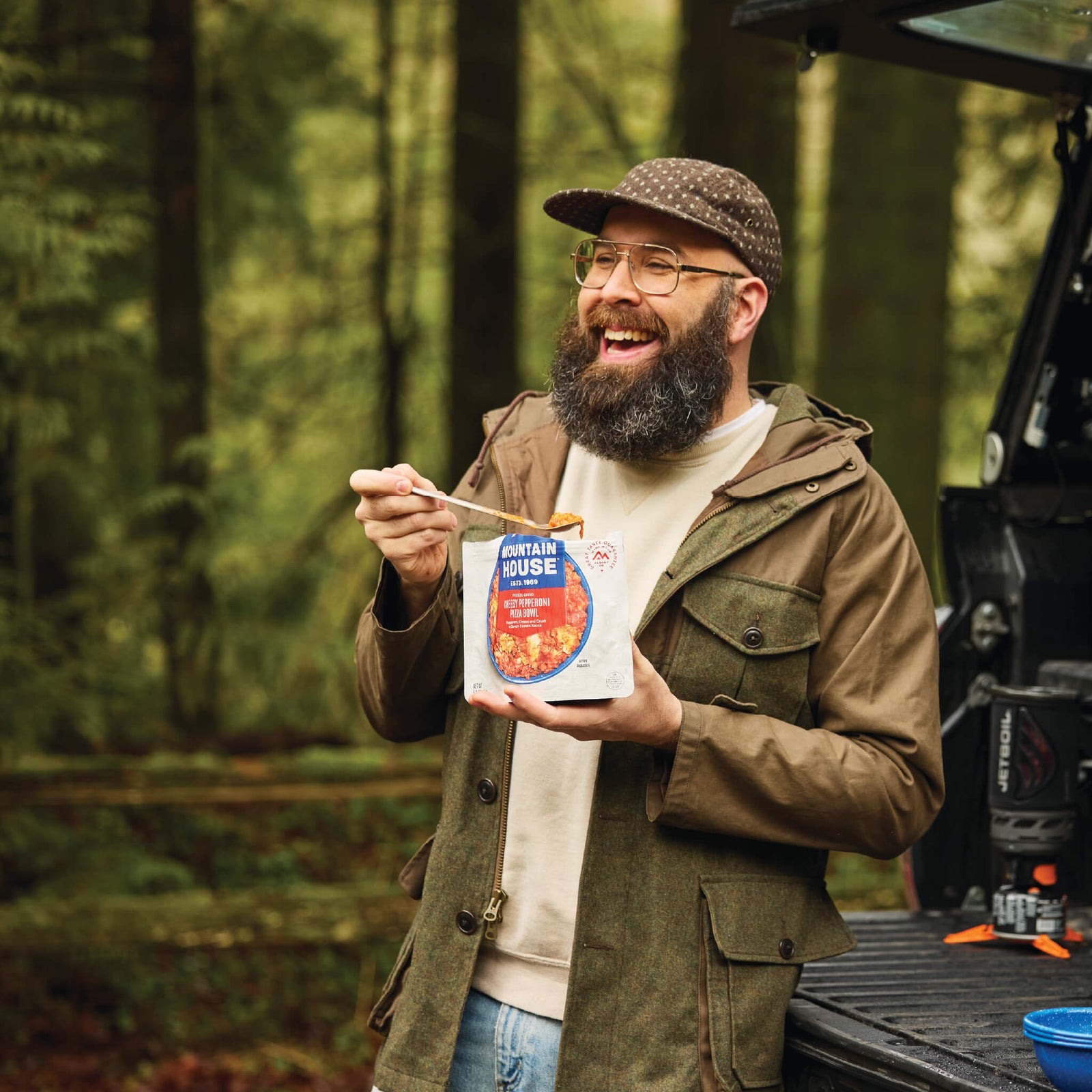 Man smiling while holding a spoonful of Mountain House Cheesy Pepperoni Pizza Bowl outside his truck in the backcountry