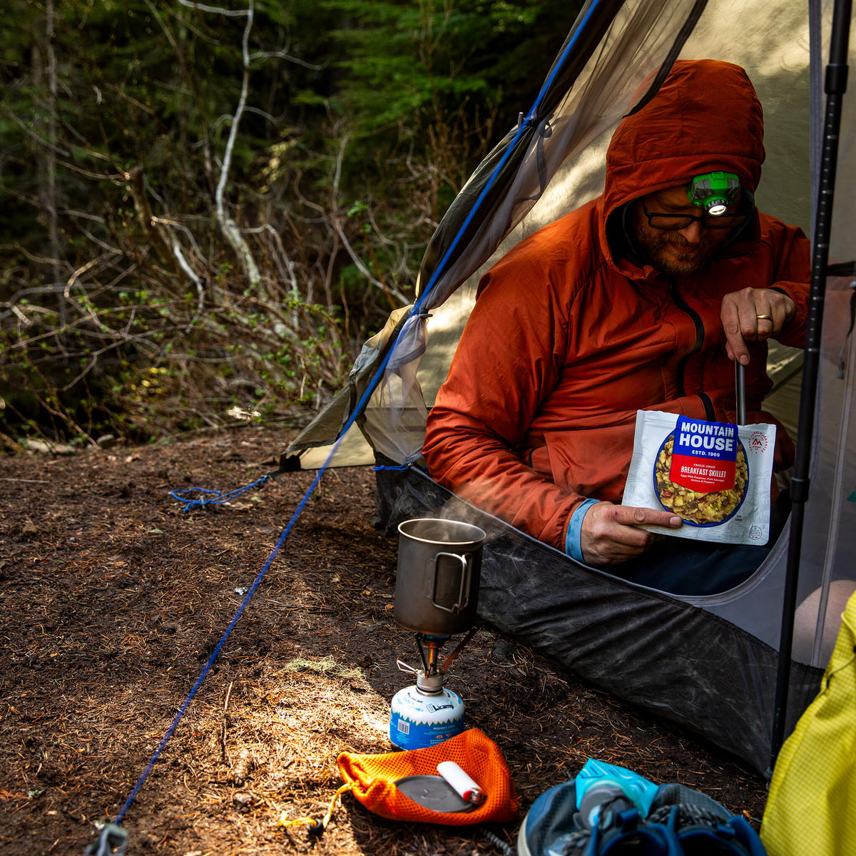Person in an orange raincoat and headlamp sits in a tent, stirring Breakfast Skillet from a pouch.