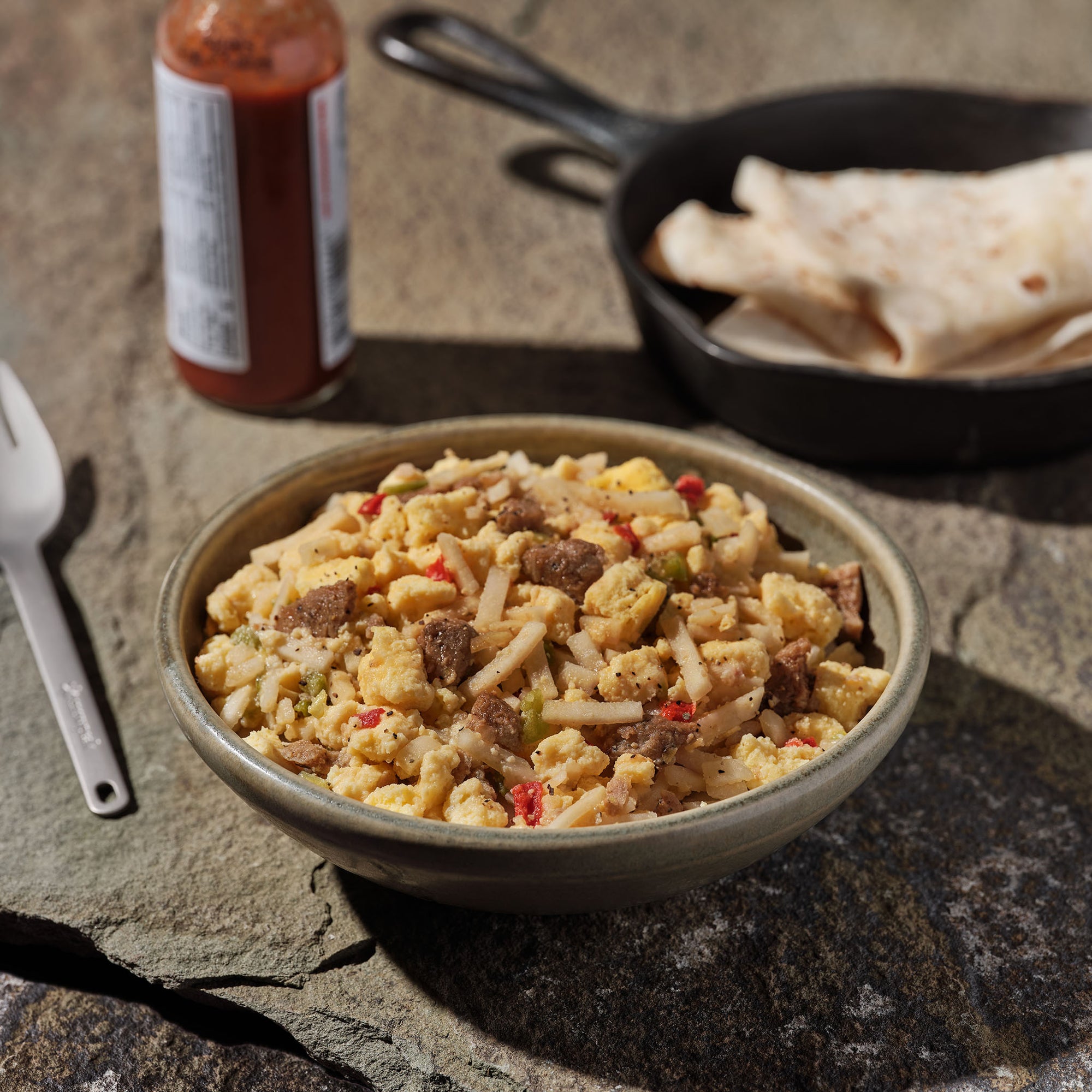 A bowl of Mountain House Breakfast Skillet on a rock surface, with a cast iron skillet of tortillas, hot sauce, and a fork behind it.