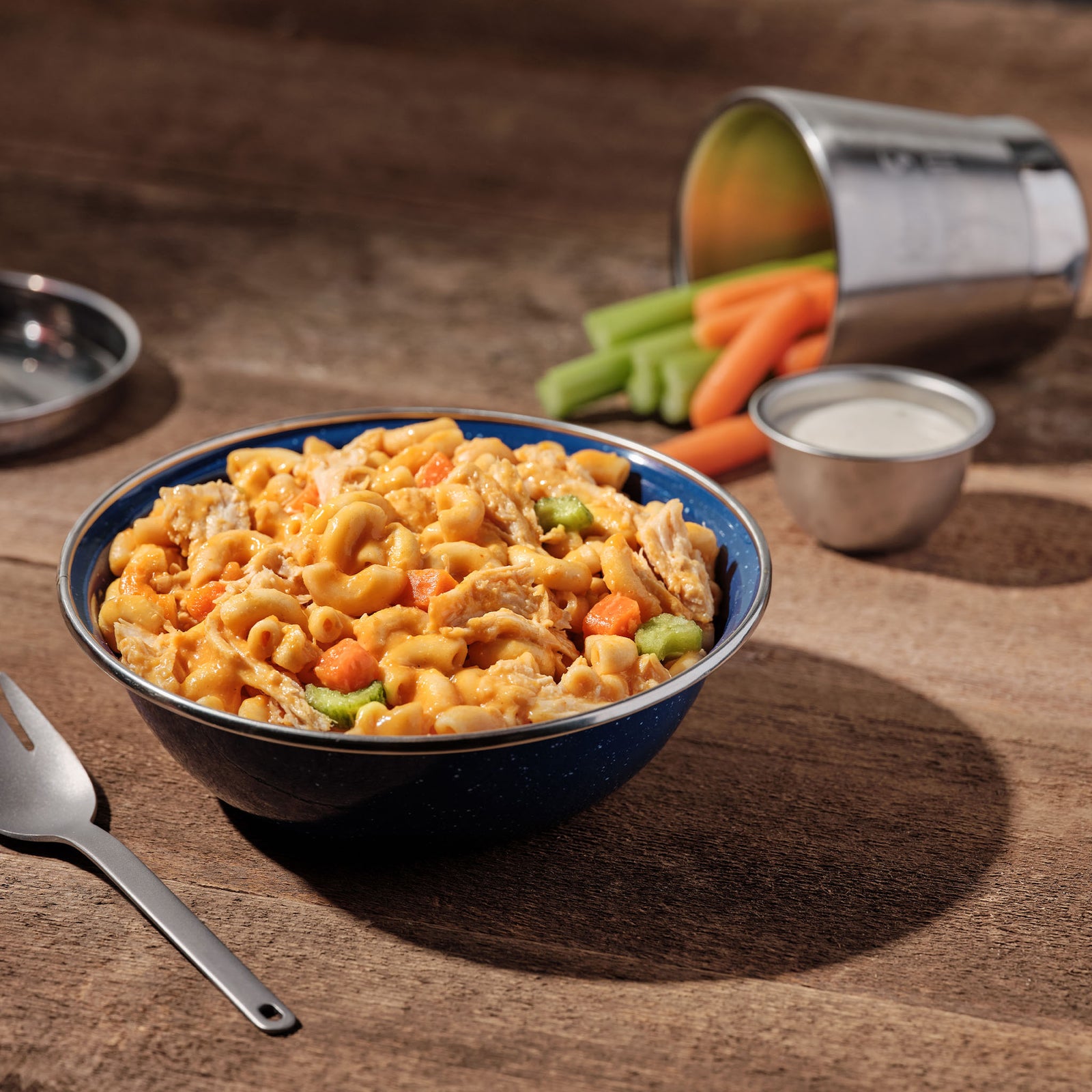 A blue camping bowl filled with Mountain House Buffalo-Style Chicken Mac & Cheese is on a picnic table, accompanied by a side of carrots and celery with ranch dressing in the background.