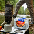 Person resting foot on a river rock with Cajun Style Jambalaya pouch and camp stove on a small table.