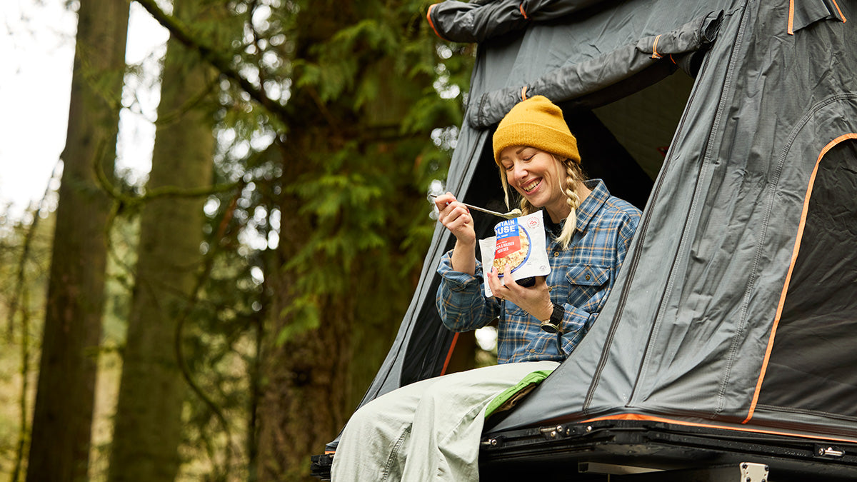 Person smiling while eating Mountain House Chicken & Mashed Potatoes from a pouch in a roof tent.