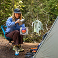Girl sitting in a camp chair beside a tent, pouring water into a Mountain House Chicken Fajita Bowl pouch.