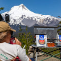 Person drinking from a can with a Mountain House pouch on a camping table and snow-capped mountain in the background.