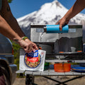 Person pours water into a Chicken Fried Rice pouch held open by another person at a camping table.