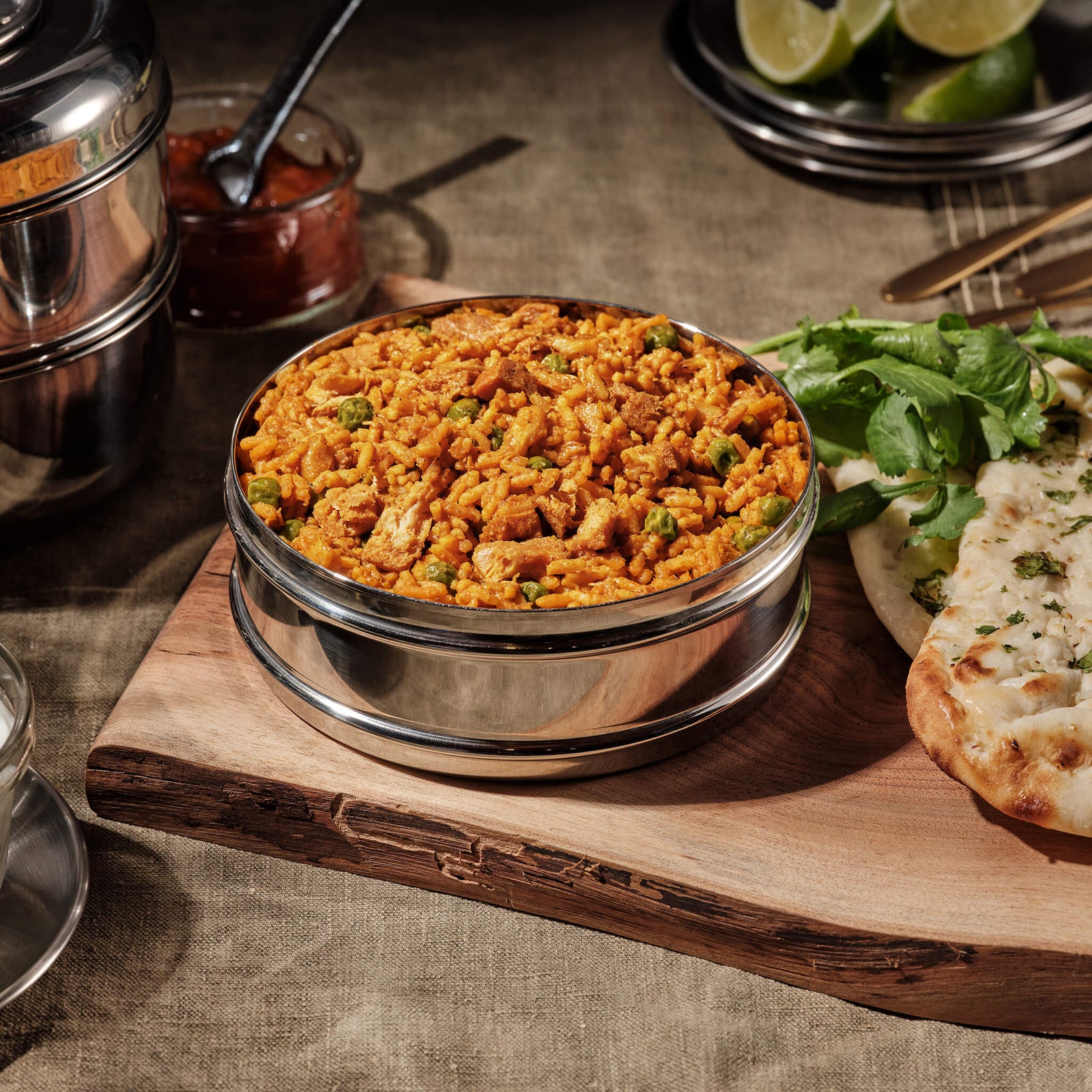 Stainless steel bowl of chicken tikka masala on a wooden board with a side of naan bread.