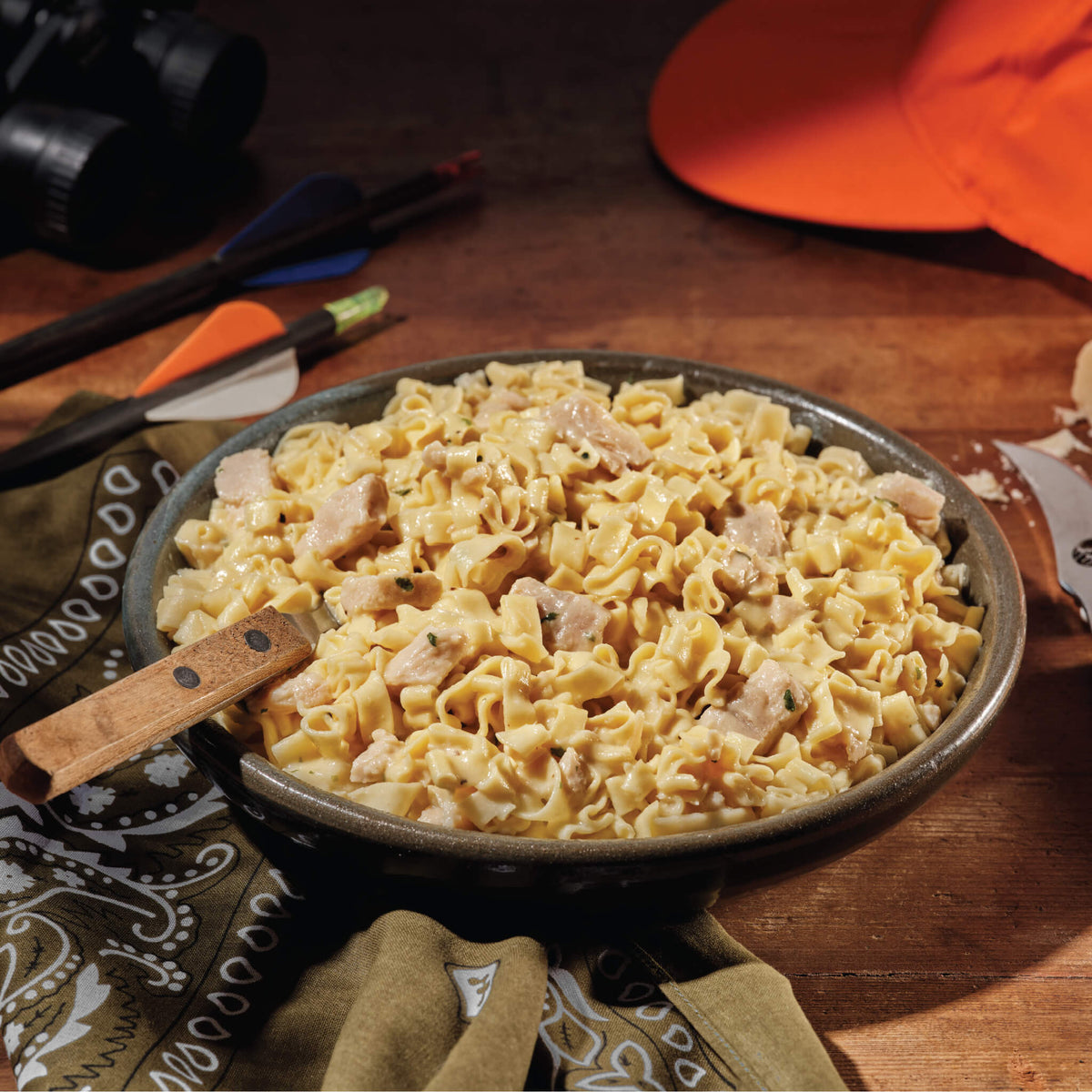 A bowl of Mountain House Fettuccine Alfredo with Chicken on a wooden countertop with hunting gear in the background.