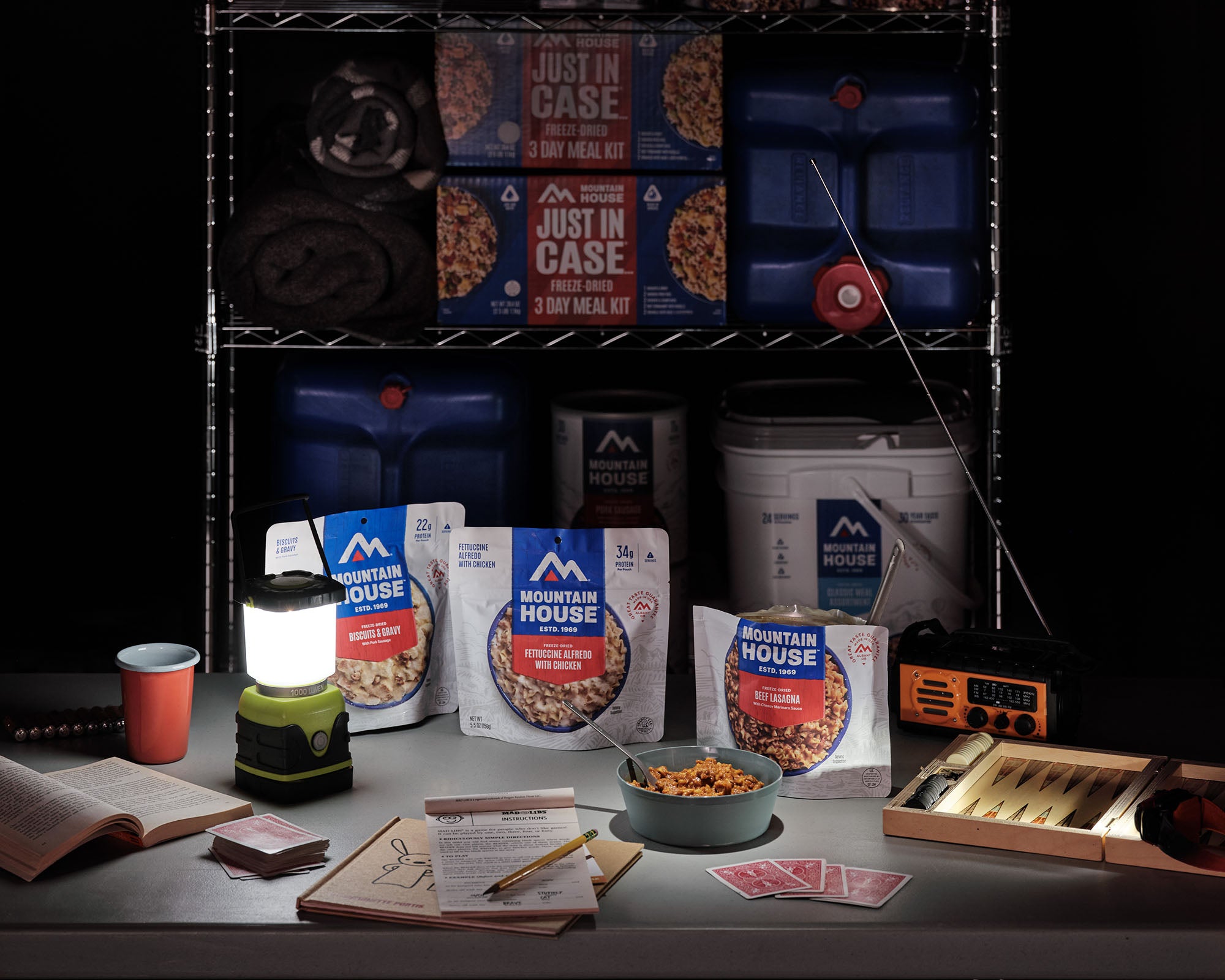 Mountain House emergency meal pouches sit on a table during a power outage alongside a weather radio, board games, playing cards, and a lantern, with additional emergency supplies on storage shelves in the background.