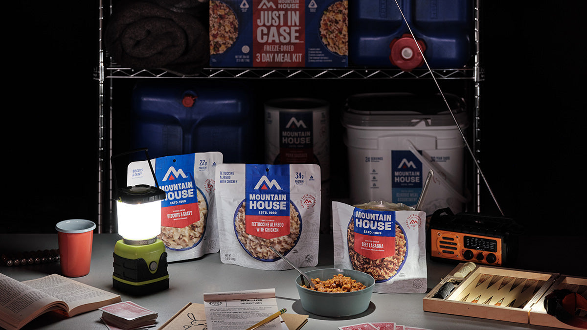 Mountain House emergency meal pouches sit on a table during a power outage alongside a weather radio, board games, playing cards, and a lantern, with additional emergency supplies visible on shelves in the background.