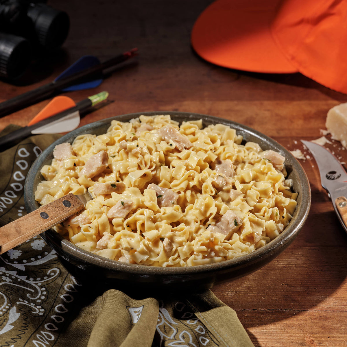 Dish of Fettuccine Alfredo with Chicken with a wooden spoon on a rustic wooden table with hunting equipment.
