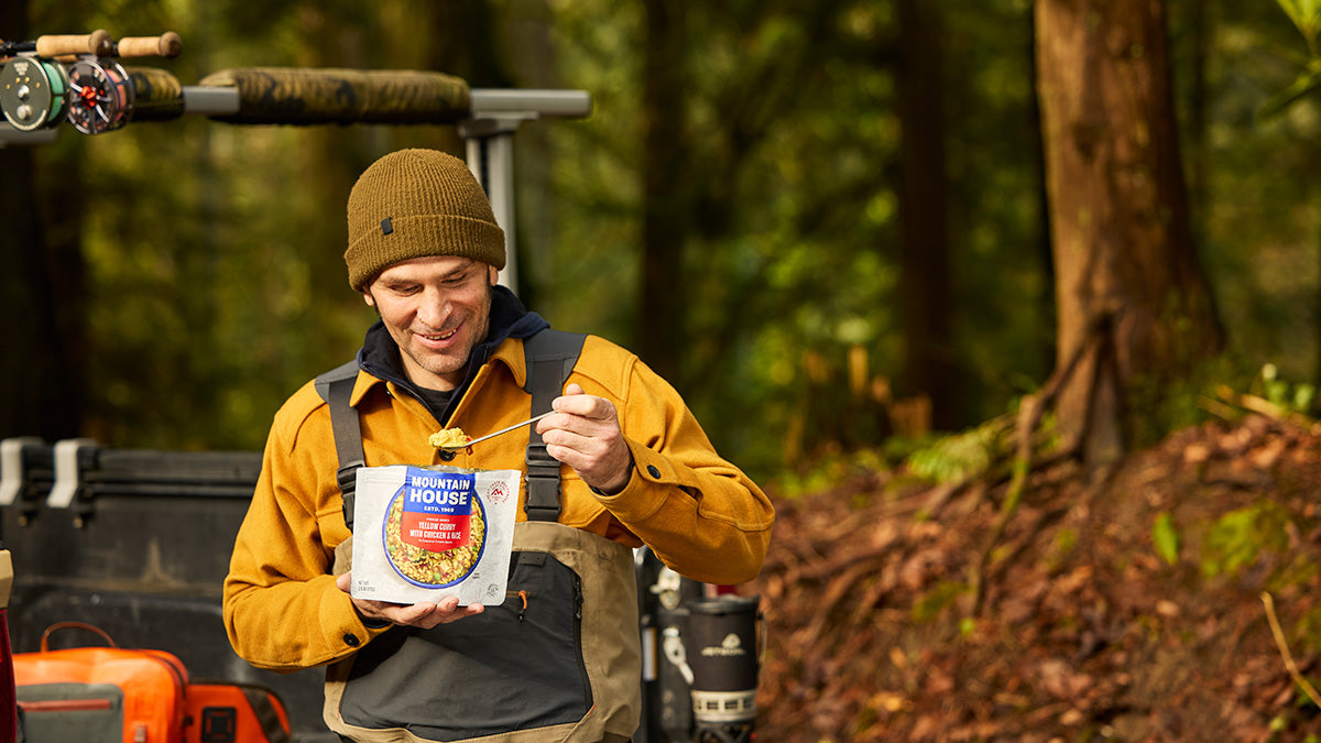 A man wearing waders stands beside a UTV during a fishing trip, eating Mountain House Yellow Curry with Chicken & Rice outdoors.