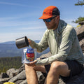 A guy pouring water into a Mountain House pouch while sitting on a boulder.