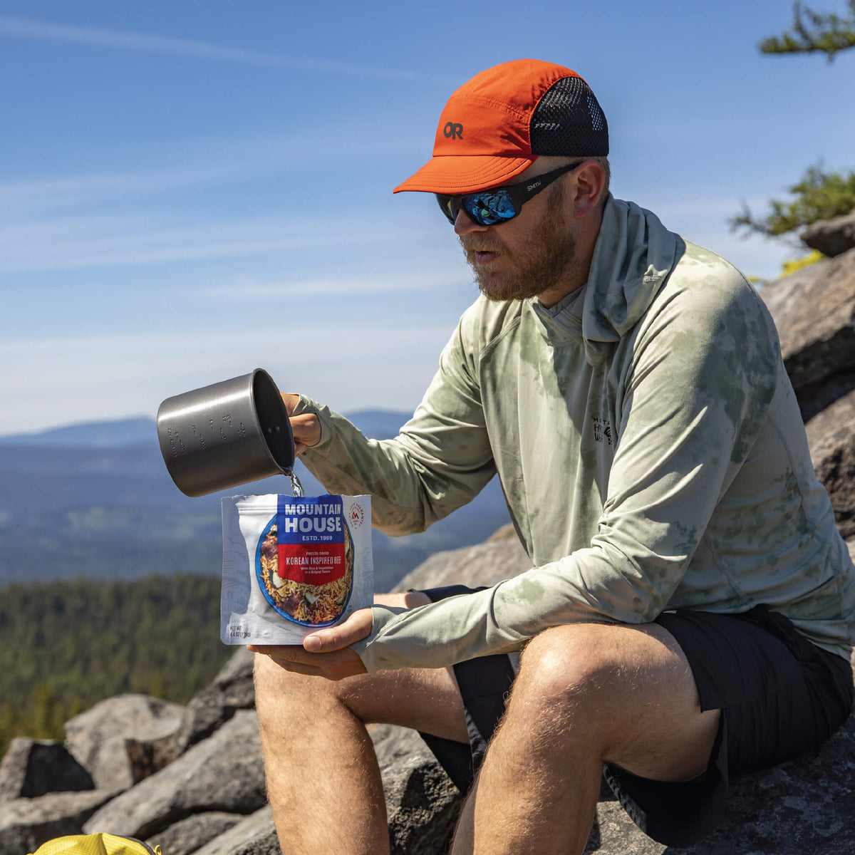 A guy pouring water into a Mountain House pouch while sitting on a boulder.