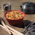 Red bowl of Kung Pao Chicken with kettle and cup in background.