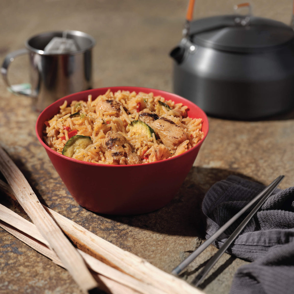 Red bowl of Kung Pao Chicken with kettle and cup in background.