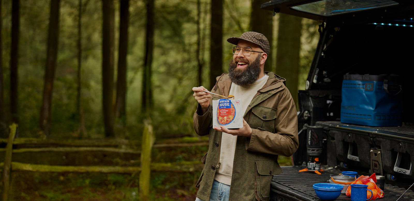 Man with a beard eating from a Mountain House pouch in front of an open truck in a forest