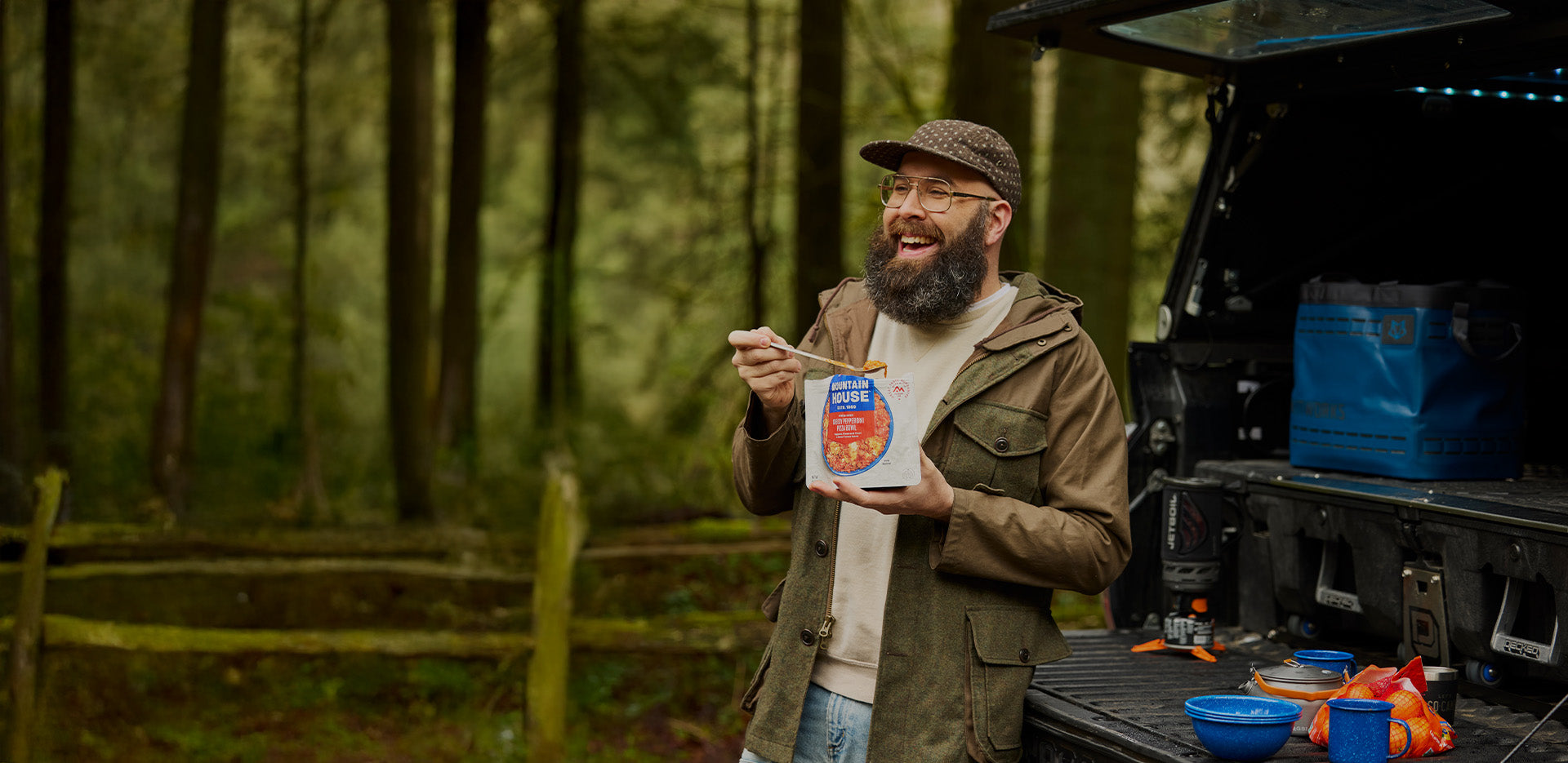 Man with a beard eating from a Mountain House pouch in front of an open truck in a forest